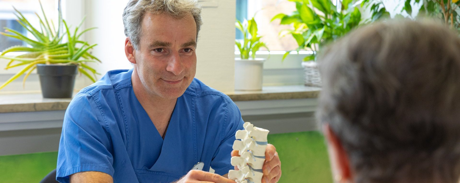 Arzt in blauer Kleidung erklärt einem Patienten mithilfe eines Wirbelsäulenmodells in einem hellen Raum mit Pflanzen.