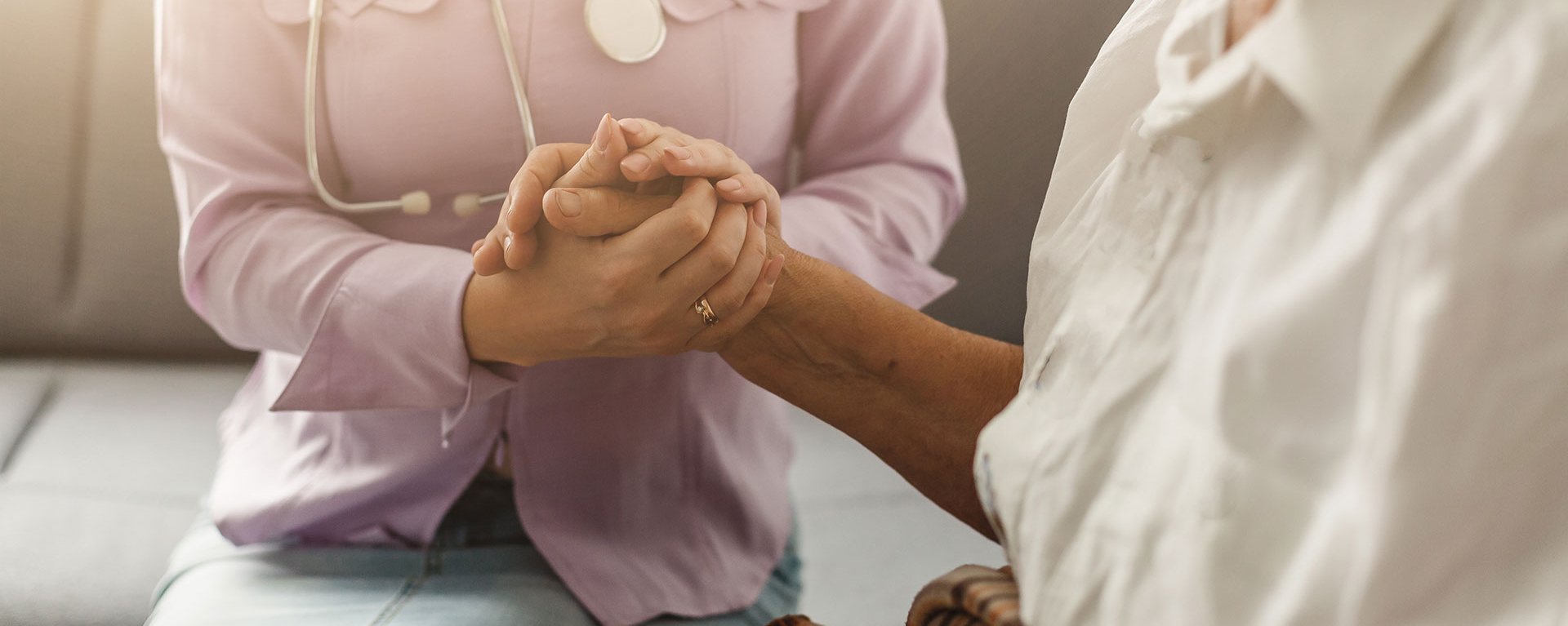 Junger Arzt des Klinikverbunds Allgäu sitzt mit einer älteren Patientin auf einer Couch und hält Ihre Hand.