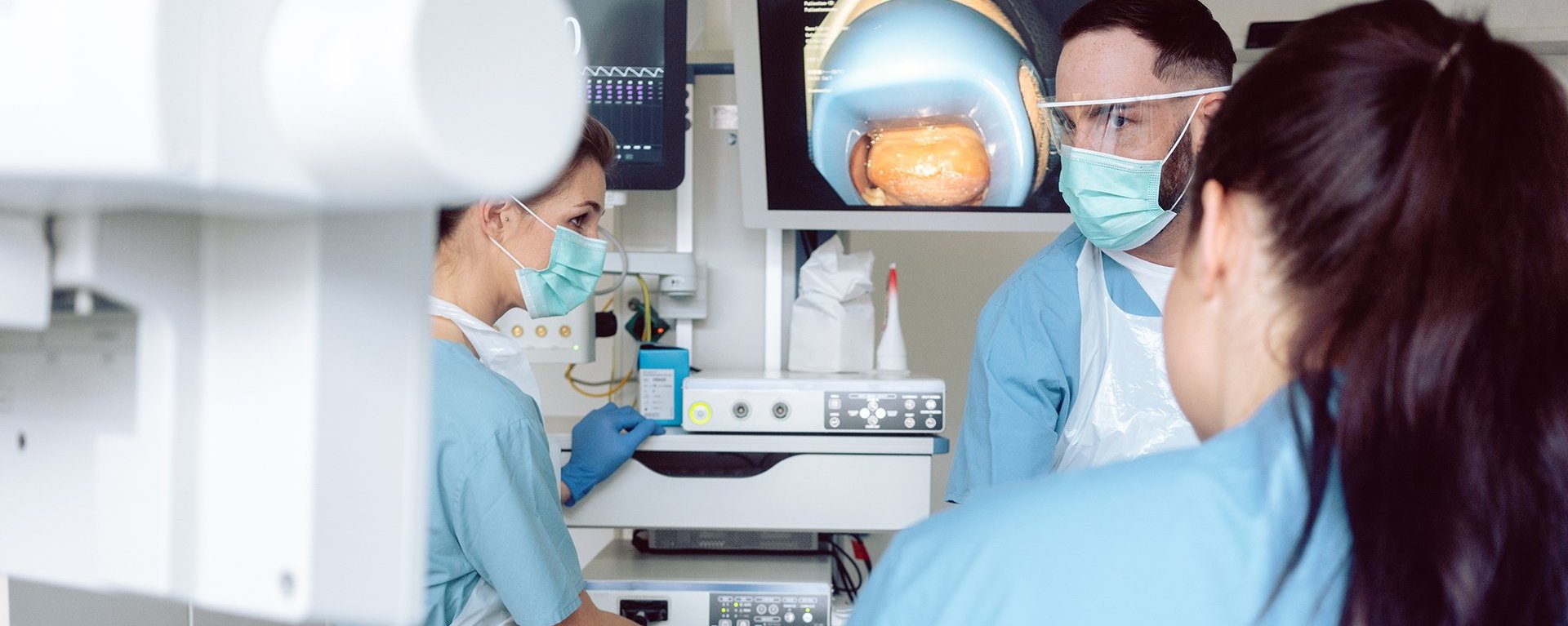 Medizinisches Team in Schutzkleidung bei einer endoskopischen Untersuchung, im Hintergrund ist auf einem Monitor das Innere eines Organs zu sehen