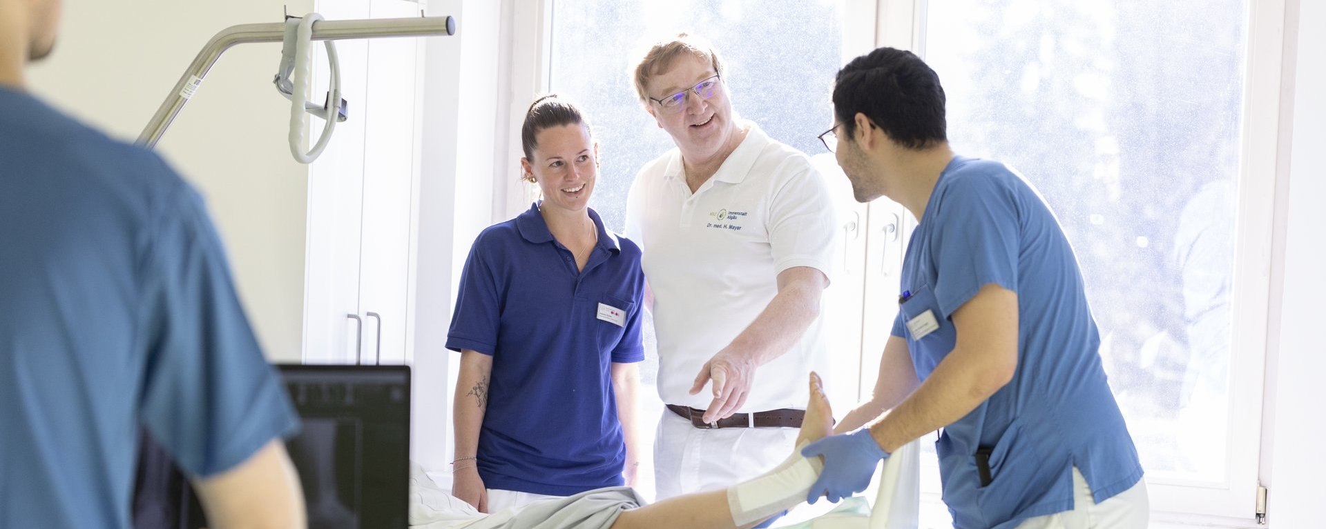 Mehrere medizinische Fachkräfte stehen gemeinsam an einem Patientenbett in einem hellen Krankenzimmer und besprechen die Behandlung, während eine Person am Bett arbeitet.