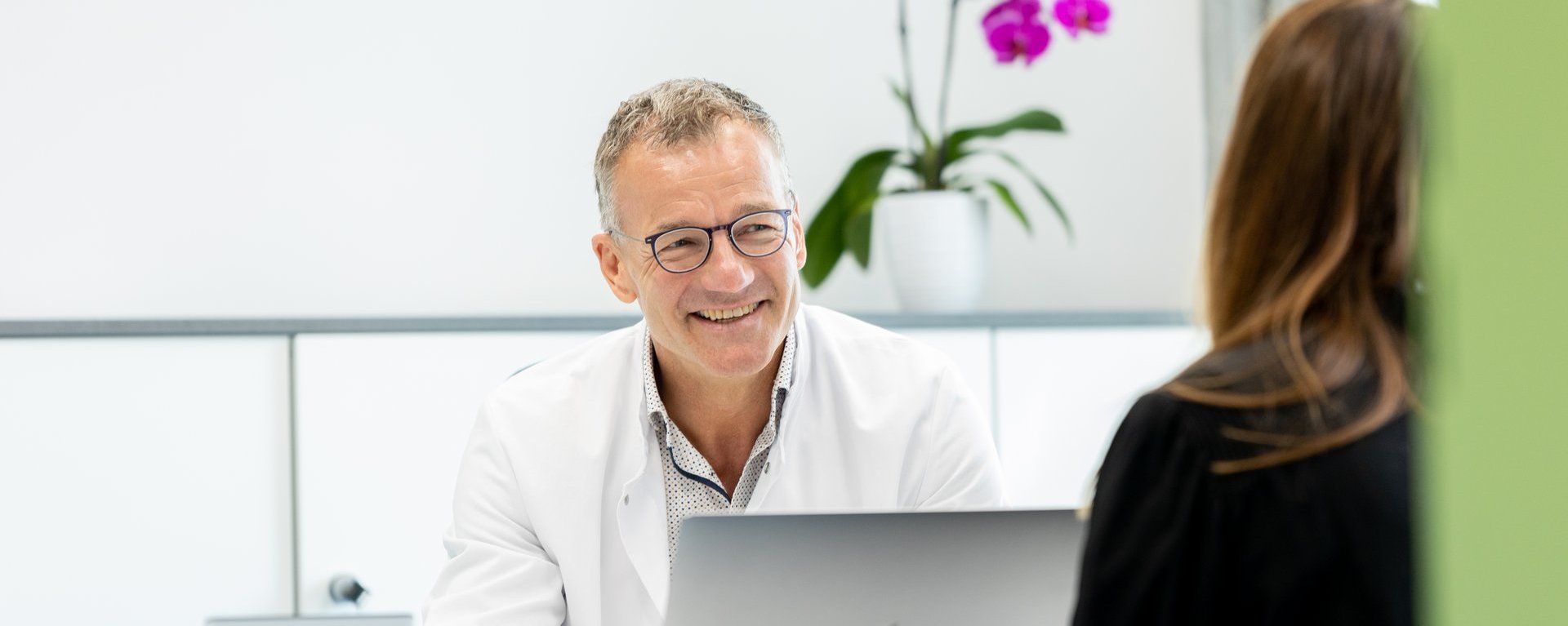 Ein Arzt im weißen Kittel sitzt an einem Schreibtisch mit Laptop und spricht mit einer Patientin; im Hintergrund steht eine violette Orchidee