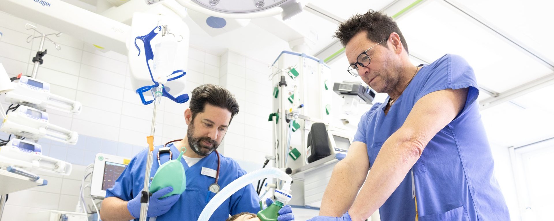 Zwei Männer beleben einen Patienten wider. Einer hält eine Sauerstoffflasche in der Hand, der andere Mann übt eine Herzdruckmassage aus.