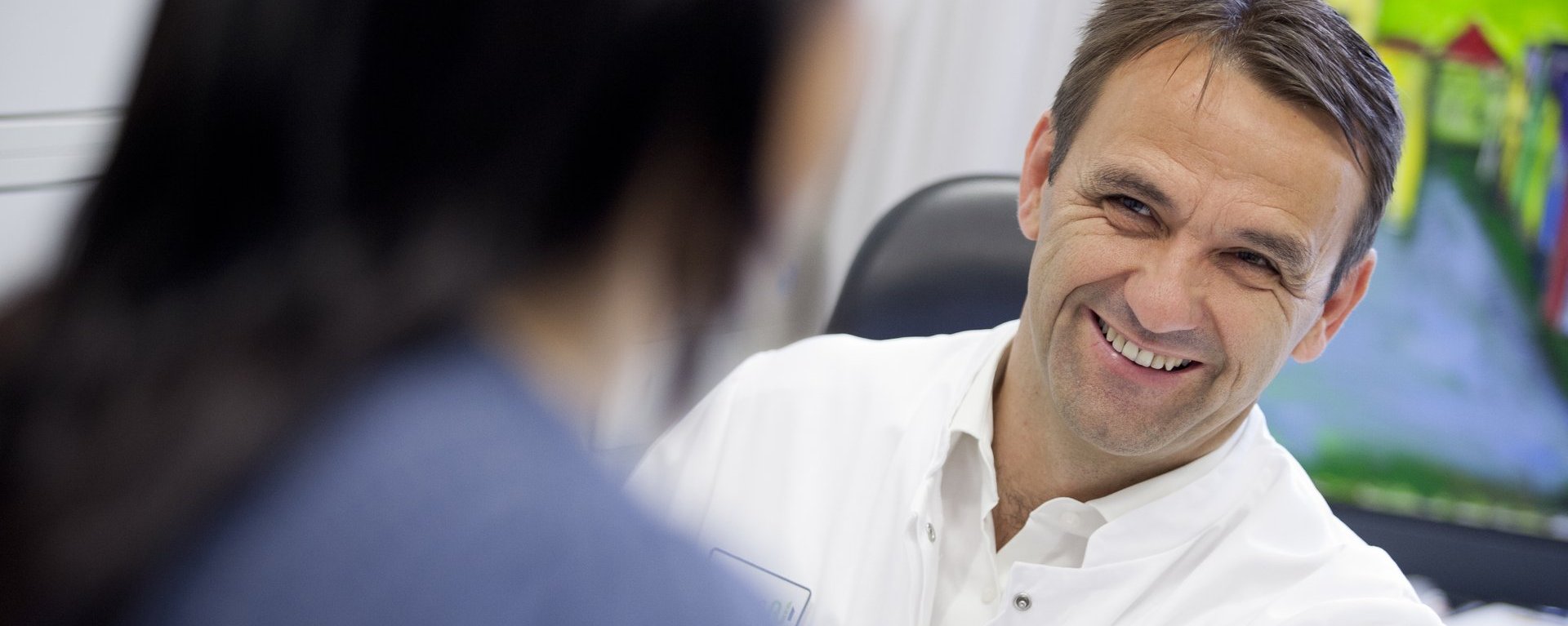 Dr. Nuscheler sitzt einer Patientin mit dunklem, langen Haar gegenüber und lächelt sie an.