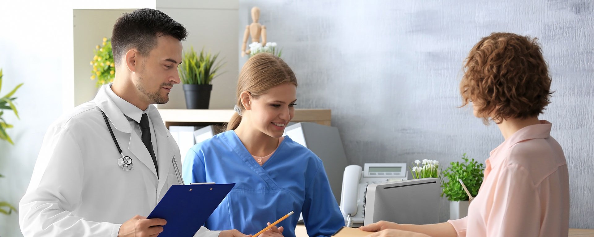 Ein Arzt und eine Medizinische Fachangestellt stehen im Klinikverbund Allgäu an der Rezeption und begrüßen eine neue Patientin.