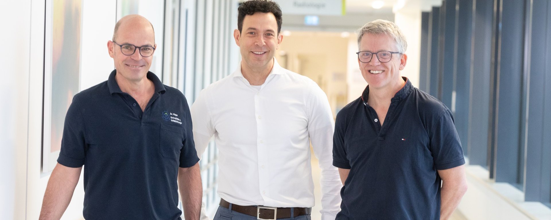 Drei Ärzte auf einem hellen Klinikflur, zwei in dunkelblauen Poloshirts und einer in weißem Hemd.