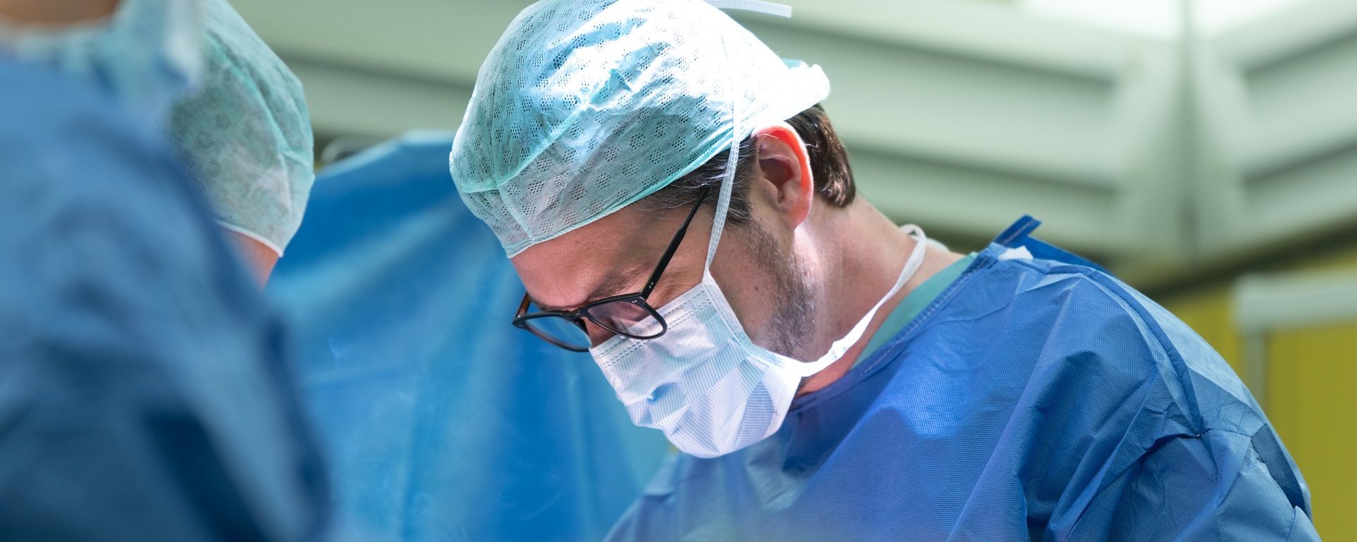 Chirurg im blauen OP-Kittel und mit Mundschutz arbeitet konzentriert während einer Operation im Operationssaal.