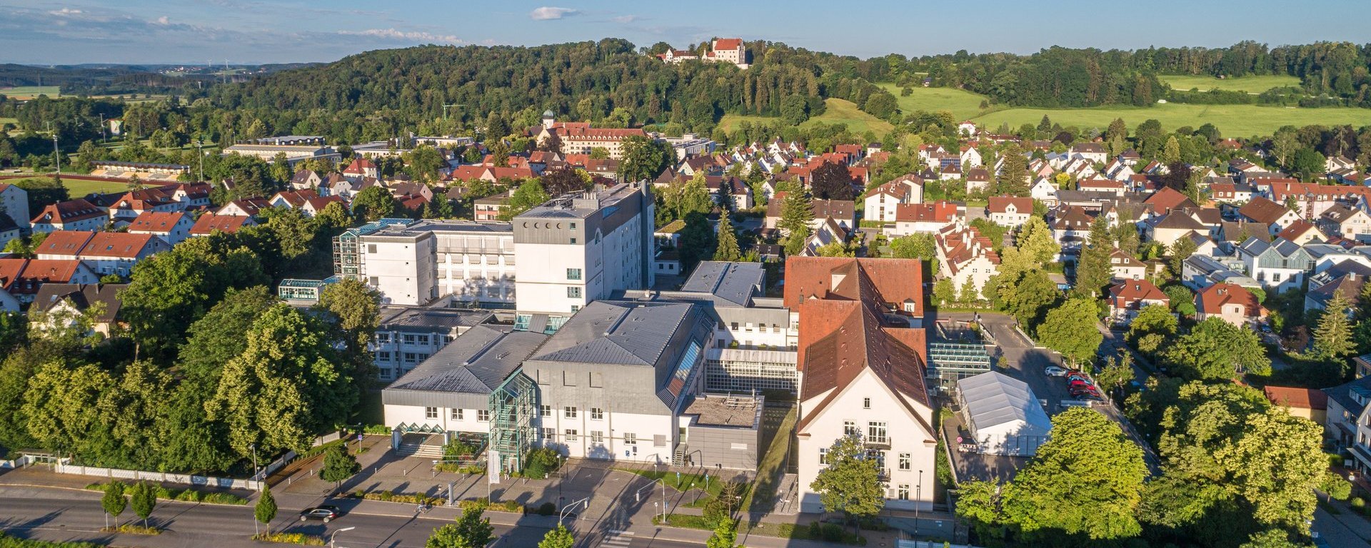 uftaufnahme der Klinik in Mindelheim, umgeben von Wohnhäusern, Bäumen und grünen Hügeln im Hintergrund bei klarem, sonnigem Wetter.