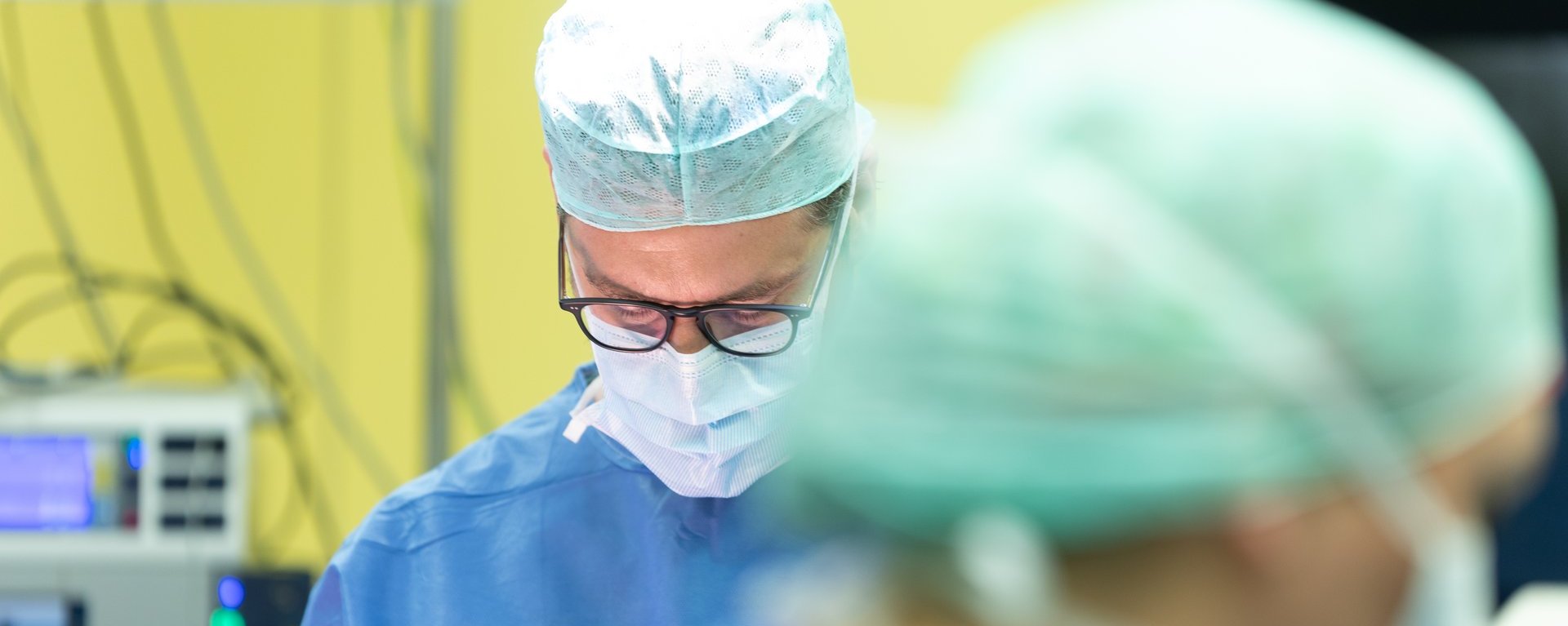 Chirurg im Operationssaal konzentriert bei einem Eingriff, trägt OP-Maske, Haube und Schutzkleidung, im Vordergrund unscharf eine weitere Person im OP-Team.