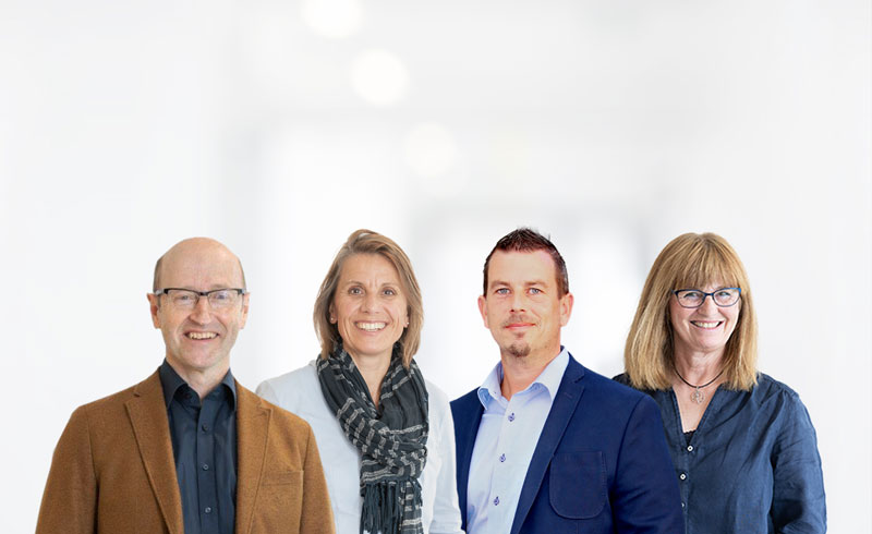 Vier Personen stehen nebeneinander vor einem hellen, unscharfen Klinik-Hintergrund: ein älterer Mann im braunen Sakko, eine Frau mit hellem Oberteil und dunklem Schal, ein Mann im blauen Sakko sowie eine Frau mit blauer Bluse. Alle blicken freundlich in die Kamera.